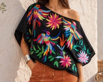May include: A black off-the-shoulder blouse featuring vibrant, multi-colored floral and bird embroidery in shades of pink, blue, yellow, and green, paired with rust-colored trousers that have visible front seam detailing.