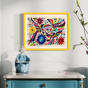 Puede incluir: Una obra de arte enmarcada con un bordado vibrante y colorido de pájaros y flores. La obra de arte está enmarcada en un marco amarillo brillante y cuelga de una pared blanca. Una figura de cerámica azul y un mueble azul están en primer plano.