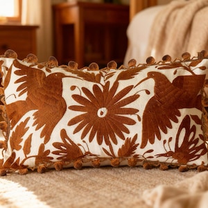 May include: Rectangular decorative pillow with a white background and brown embroidered floral and bird designs. The pillow is trimmed with brown pom-poms. The pillow is resting on a beige rug.
