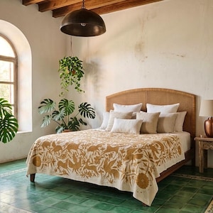 May include: A bed featuring a cream-colored quilt with a mustard yellow botanical and animal print pattern. The bed has a woven headboard and is layered with multiple neutral-toned throw pillows. A lamp sits on a wooden side table nearby.