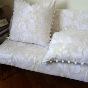 Pode incluir: Uma capa de sofá bege claro e duas almofadas quadradas a condizer, todas com um intrincado padrão bordado a branco de flores e pássaros. Cada almofada tem um acabamento com uma borda decorativa de pompons brancos nas extremidades.