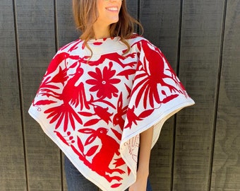May include: A white poncho with red embroidery featuring birds, flowers, and rabbits. The poncho has a square neckline and a triangular shape.