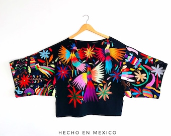May include: Black blouse with colorful embroidered birds and flowers. The embroidery features a variety of colors, including red, yellow, blue, green, and purple. The blouse has a relaxed fit and a crew neckline. The text "HECHO EN MEXICO" is printed on the bottom of the blouse.