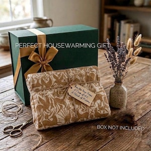 May include: A dark green gift box with a gold ribbon, a folded textile featuring a tan animal and foliage pattern, a small tag with handwritten text, a ceramic vase with dried lavender and wheat, and metal scissors on a wooden surface.