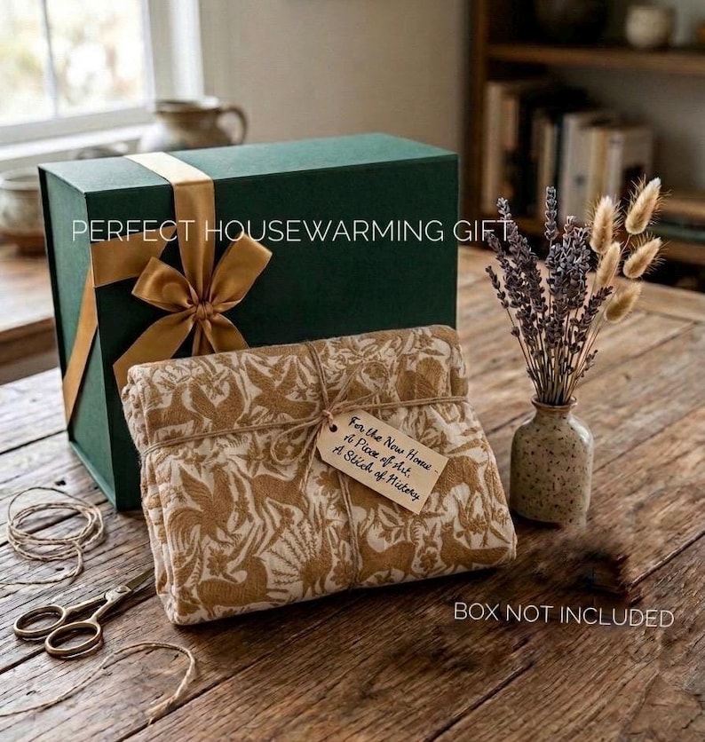 May include: A dark green gift box with a gold ribbon, a folded textile featuring a tan animal and foliage pattern, a small tag with handwritten text, a ceramic vase with dried lavender and wheat, and metal scissors on a wooden surface.