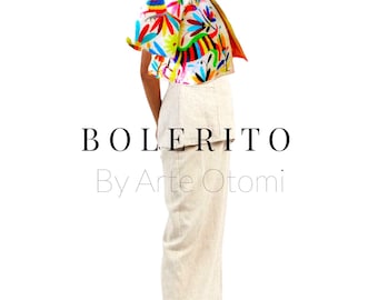 Puede incluir: Una mujer lleva una blusa blanca con un estampado floral colorido y una falda blanca. La blusa tiene un cuello con volantes y la falda es una falda larga y recta. La mujer lleva sandalias amarillas. El texto "BOLERITO By Arte Otomi" está impreso debajo de la mujer.