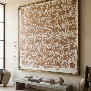 May include: A large rectangular wall tapestry featuring a tan-colored intricate pattern of various animals and floral motifs on a cream-colored fabric background. The piece is encased in a thin gold-toned frame and measures 74 inches width by 78 inches height.