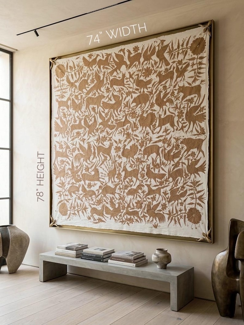 May include: A large rectangular wall tapestry featuring a tan-colored intricate pattern of various animals and floral motifs on a cream-colored fabric background. The piece is encased in a thin gold-toned frame and measures 74 inches width by 78 inches height.