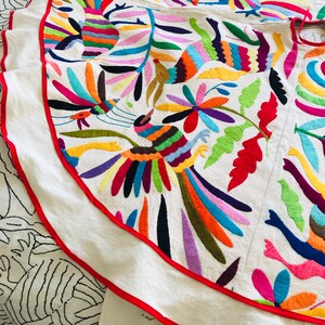 Pode incluir: Uma saia de tecido branco com bordados vibrantes e multicoloridos de p&aacute;ssaros, flores e folhagens. A pe&ccedil;a circular &eacute; finalizada com um debrum vermelho brilhante ao longo da bainha curva, exibindo um trabalho de agulha intrincado em tons de azul, laranja, rosa, amarelo e verde.