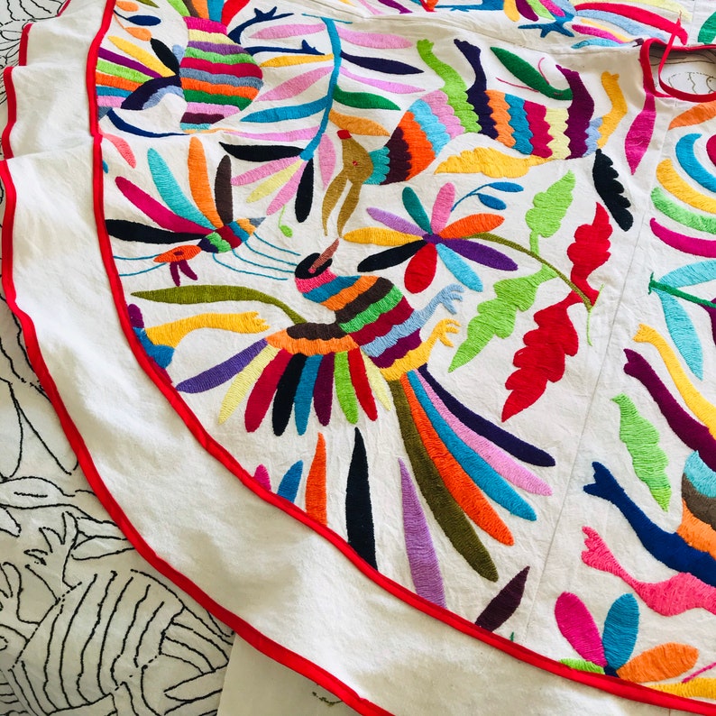 Pode incluir: Uma saia de tecido branco com bordados vibrantes e multicoloridos de p&aacute;ssaros, flores e folhagens. A pe&ccedil;a circular &eacute; finalizada com um debrum vermelho brilhante ao longo da bainha curva, exibindo um trabalho de agulha intrincado em tons de azul, laranja, rosa, amarelo e verde.