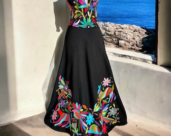 May include: A black strapless dress with a colorful embroidered bodice and skirt. The skirt has a floral and bird design. The dress is displayed on a mannequin against a white wall with a window view of the ocean.