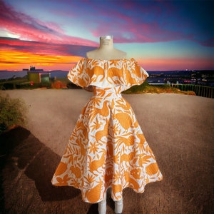May include: An off-the-shoulder dress with a white background and an orange floral and animal pattern. The dress is displayed on a mannequin. The dress has a ruffled top and a flared skirt. The background features a sunset over a landscape.