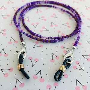 Puede incluir: Una cadena para gafas hecha de cuentas con una mezcla de semillas de color p&uacute;rpura, lavanda y violeta oscuro. La cadena est&aacute; terminada con cierres de langosta plateados, espirales de metal dorado y bucles de goma negra.