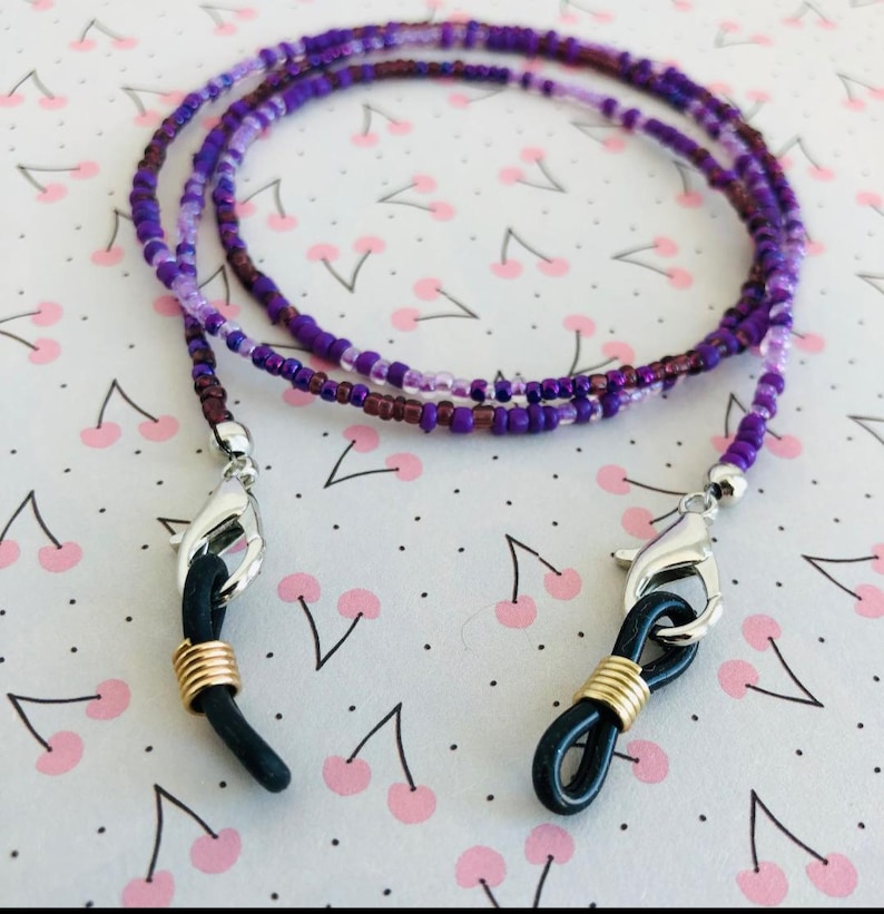Puede incluir: Una cadena para gafas hecha de cuentas con una mezcla de semillas de color p&uacute;rpura, lavanda y violeta oscuro. La cadena est&aacute; terminada con cierres de langosta plateados, espirales de metal dorado y bucles de goma negra.
