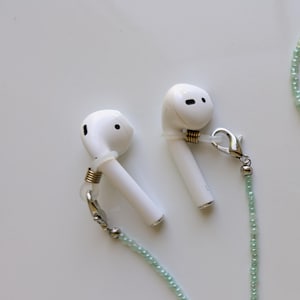 Puede incluir: Un par de auriculares inal&aacute;mbricos blancos unidos a una cadena de cuentas de color verde claro. Cada auricular tiene un lazo de silicona transparente asegurado con una peque&ntilde;a bobina de metal dorado y un cierre de mosquet&oacute;n plateado.