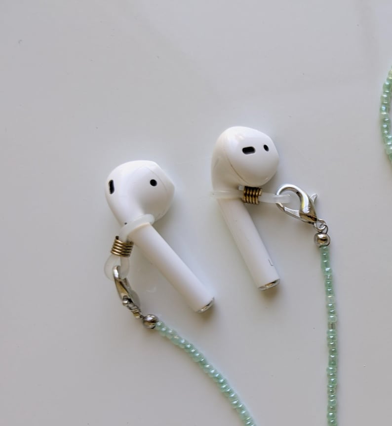 Puede incluir: Un par de auriculares inal&aacute;mbricos blancos unidos a una cadena de cuentas de color verde claro. Cada auricular tiene un lazo de silicona transparente asegurado con una peque&ntilde;a bobina de metal dorado y un cierre de mosquet&oacute;n plateado.