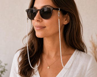 Puede incluir: Unas gafas de sol de color marrón oscuro con una cadena de cuentas de perlas blancas unida a las patillas. La cadena presenta pequeñas cuentas blancas uniformes y se conecta a las gafas con cierres metálicos plateados y bucles de silicona.