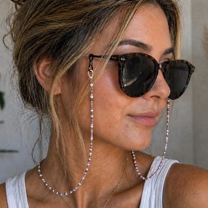 May include: A beaded spectacle chain featuring small, multi-coloured beads in shades of pink, white, and dark grey. The chain attaches to the temples of dark brown tortoiseshell sunglasses with silver-toned metal clasps and black rubber loops.