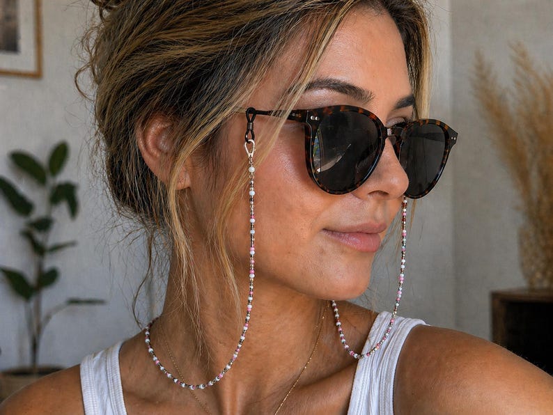 Puede incluir: Una cadena para gafas con peque&ntilde;as cuentas multicolores en tonos rosa, blanco, gris y negro. La cadena se sujeta a las patillas de unas gafas de sol de carey marr&oacute;n oscuro mediante cierres de metal plateado y bucles de goma negros.