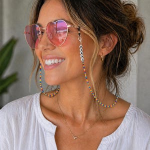 May include: Heart-shaped sunglasses with a gold frame and pink lenses. The glasses are connected to a beaded chain with the name "SARAH" spelled out in white letters. The chain features a rainbow of colours.