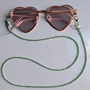May include: Heart-shaped sunglasses with a rose gold frame and pink lenses. The glasses are attached to a beaded eyeglass chain. The chain is made of small, light green beads with gold accents.