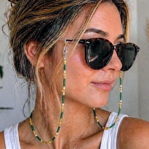 May include: Sunglasses with a beaded chain in green, gold, and white. The chain is attached to the sunglasses and hangs down. The sunglasses have a dark tortoiseshell frame and dark lenses. The person is wearing a white vest top.