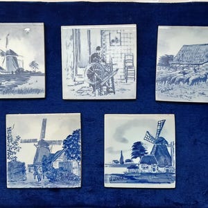 May include: Five blue and white ceramic tiles with Dutch scenes. Each tile features a different illustration, including windmills, a barn, and figures working. The tiles are arranged on a dark blue surface.