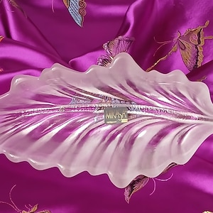 May include: A clear, frosted glass leaf-shaped serving dish. The dish has a textured surface and a wavy edge. The Mikasa Germany label is visible. The dish is set against a vibrant purple background.