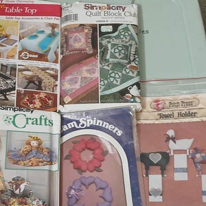 May include: A collection of Simplicity craft pattern packets, including table top accessories, quilt block club, and craft projects. The packets feature images of table settings, quilts, and craft projects. A "Towel Holder" packet is also visible.