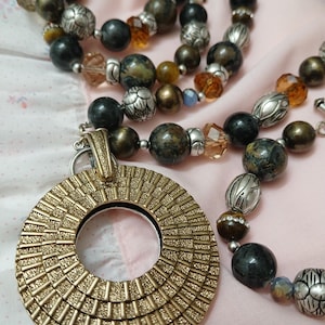 May include: A gold-colored pendant necklace with a large circular pendant and beaded necklace strands. The necklace features a variety of beads in shades of brown, silver, and clear. The pendant has a textured design.