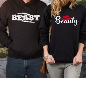 May include: Two black hoodies with white text. One hoodie says "BEAST" in a bold font with a white claw mark design. The other hoodie says "Beauty" in a cursive font with a red lip design.