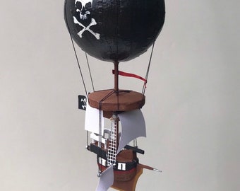 Può includere: Un pallone aerostatico nero con un designo di teschio e ossa incrociate bianche. Il pallone è attaccato a una piccola piattaforma di legno con una vela bianca e una bandiera rossa.