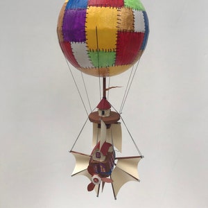 May include: A colorful patchwork hot air balloon with a small house attached to the bottom. The house has a red roof and white walls with windows. The balloon is suspended by strings.