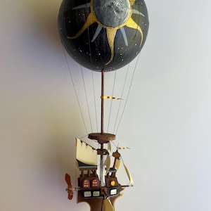 Op de afbeelding: Handgemaakte luchtballonsculptuur met een hemels thema. De zwarte bol is versierd met een zon- en maanontwerp. De gondel eronder heeft een scheepsvormig ontwerp met zeilen en een propeller. De sculptuur wordt opgehangen door dunne draden.