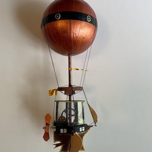 May include: A handcrafted hot air balloon model with a copper-coloured spherical top, a black band, and a detailed gondola. The gondola features a propeller, sails, and a transparent enclosure with gears. The balloon is suspended by thin wires.