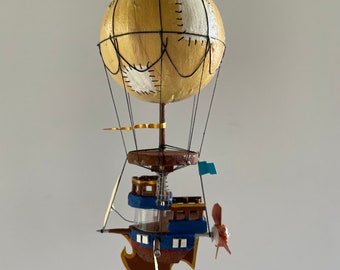 Può includere: Un modello fatto a mano di una mongolfiera con una nave attaccata alla parte inferiore. Il pallone è fatto di carta e ha un design oro e bianco. La nave è fatta di legno e ha uno schema di vernice blu e marrone.