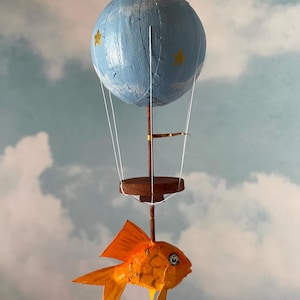 Puede incluir: Un globo aerostático de papel maché azul con estrellas doradas y un pequeño pez naranja de papel maché colgando de la parte inferior.