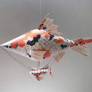 Op de afbeelding: Een wit, oranje en zwart papier-maché vis-luchtschip met een kleine gondel. De vis heeft een lange, puntige snuit en een grote, waaiervormige staart.