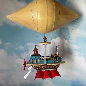 May include: A whimsical, handcrafted model of a dirigible airship with a blue and green ship attached to a yellow balloon. The ship has red sails and a propeller.