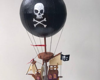 Può includere: Un pallone aerostatico nero con un disegno di teschio e ossa incrociate bianche. Il pallone è attaccato a una nave pirata marrone con vele bianche e una bandiera pirata nera.