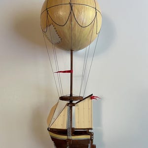 Könnte beinhalten: Eine handgefertigte Skulptur, die einen Heißluftballon und ein Segelboot kombiniert. Der Ballon ist eine hellbeige Kugel mit schwarzen Details. Das Segelboot ist braun mit beigen Segeln und roten Flaggen, die mit Drähten unter dem Ballon hängen. Ein einzigartiges Dekorationsstück.