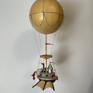 May include: A handcrafted hot air balloon model with a tan balloon, black lines, and a detailed gondola. The gondola features a propeller, a silver structure, and small red flags. The balloon is suspended by thin black strings.