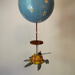May include: A blue hot air balloon with white daisy flowers painted on it is attached to a wooden platform. A small wooden turtle with a yellow shell and a figure on top is attached to the platform. The turtle is holding a fishing rod.