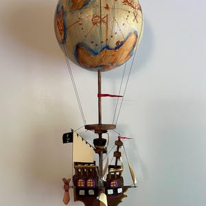 Peut inclure: Une sculpture en bois artisanale d'une montgolfière avec un globe et une base de bateau pirate. Le globe est peint avec un motif de carte dans des tons de marron, d'or et de bleu. Le bateau pirate présente des voiles, un drapeau noir et des détails complexes.