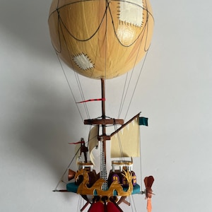 May include: A handcrafted hot air balloon model. The balloon is light tan with black lines and white patches. The gondola has a teal base, gold accents, and a red wing. A propeller and small sails complete the design.
