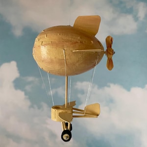 May include: A handmade model of a blimp with a propeller and a small plane attached to the bottom. The blimp is made of paper and painted a light brown colour. The plane is also made of paper and painted a light brown colour. The model is hanging from the ceiling against a blue sky with white clouds.