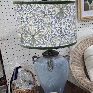 May include: A blue ceramic lamp with a floral patterned lampshade. The lampshade features a white background with blue and green floral designs and a green trim. A small wicker basket with a blue ribbon sits to the left of the lamp.