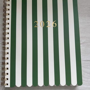 May include: A spiral-bound planner with a green and white striped cover. The cover features the gold text "2026". The planner has a scalloped edge and a gold spiral binding.