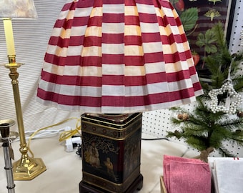 May include: A table lamp featuring a pleated lampshade with horizontal red and white stripes. The base is a rectangular black metal tin decorated with gold-colored Asian-inspired illustrations, mounted on a dark wooden square stand.