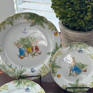 May include: A set of three white ceramic plates featuring illustrations of Peter Rabbit. The plates have a scalloped edge with a green and yellow floral border. The plates are displayed on black metal stands.
